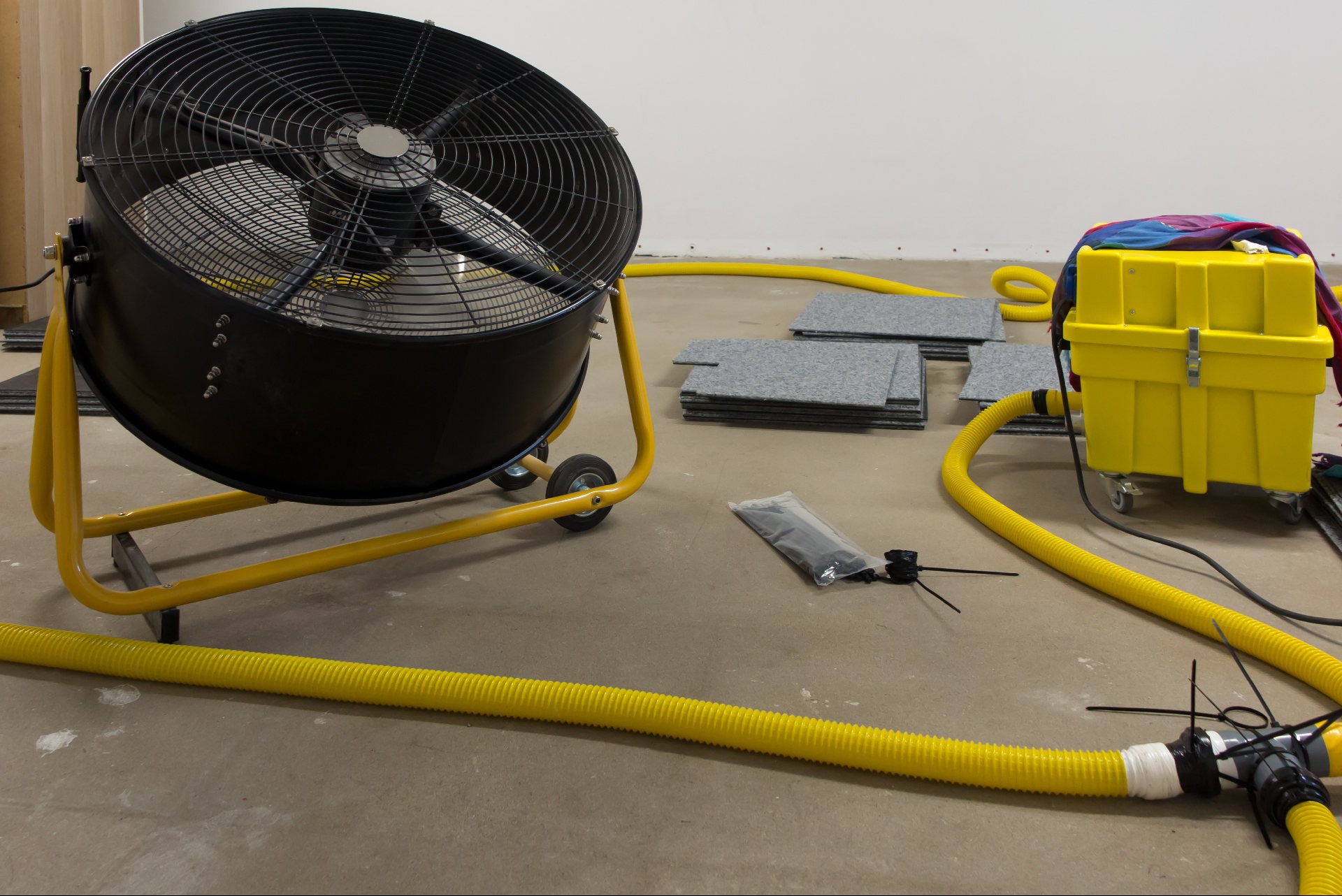 Profesjonalny wentylator osuszający z żółtymi wężami i akcesoriami na szarym podłożu. Widoczne płytki podłogowe i żółty pojemnik. Sprzęt do usuwania wilgoci.