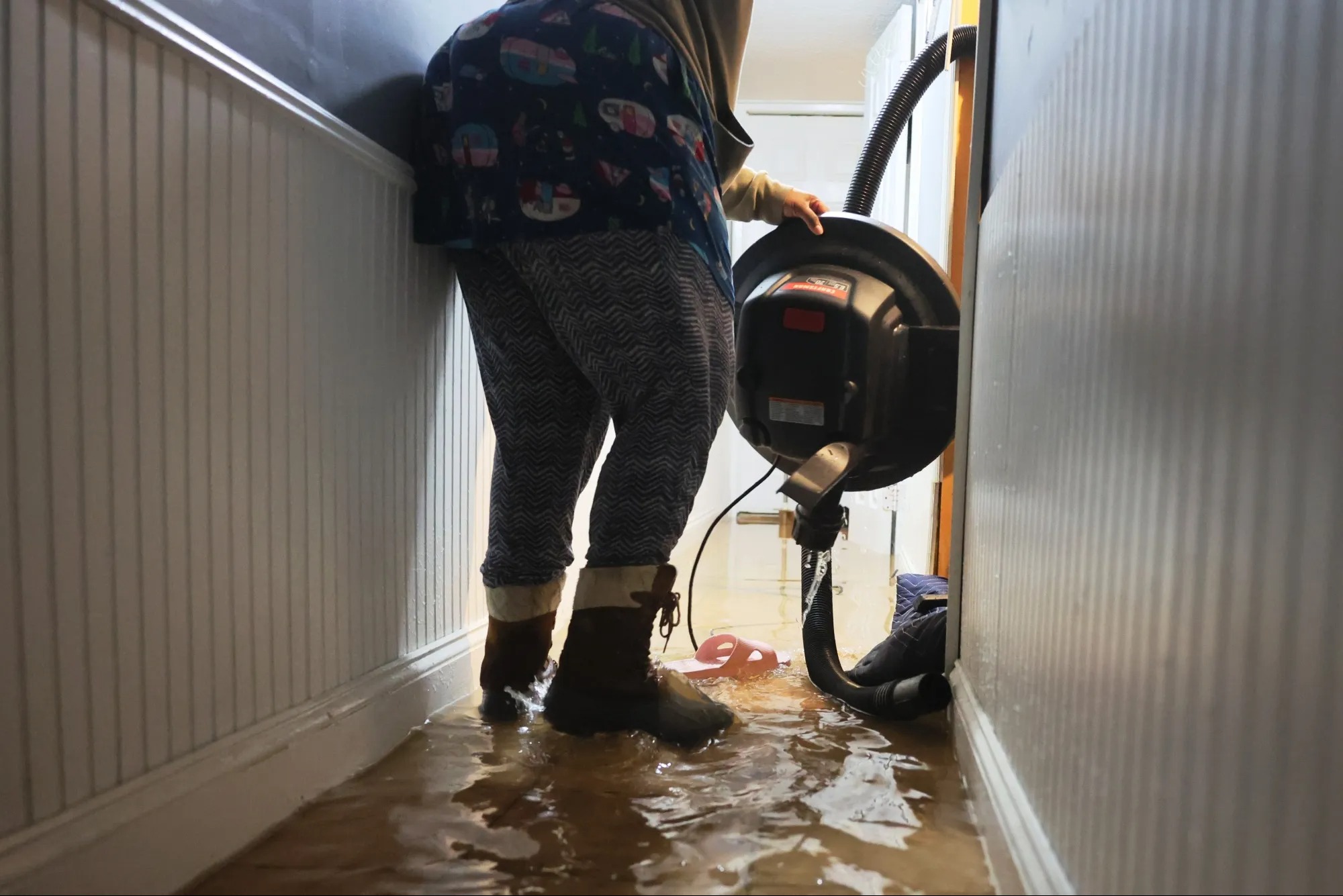 Zalana podłoga w korytarzu, osoba w kaloszach używa przemysłowego odkurzacza do usunięcia wody. Widoczne panele ścienne i fragment otwartych drzwi.