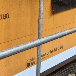 Dom szkieletowy - Fragment ściany domu w budowie, z widoczną izolacją 'naturheld 180', szarą rurą rusztowania i fragmentem czarnej membrany.