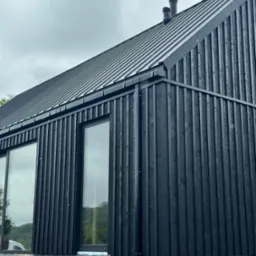 Czarny dom szkieletowy z metalowym dachem i dużymi oknami, wykończony pionowymi deskami. Widoczny fragment elewacji i otoczenie z kamieni.