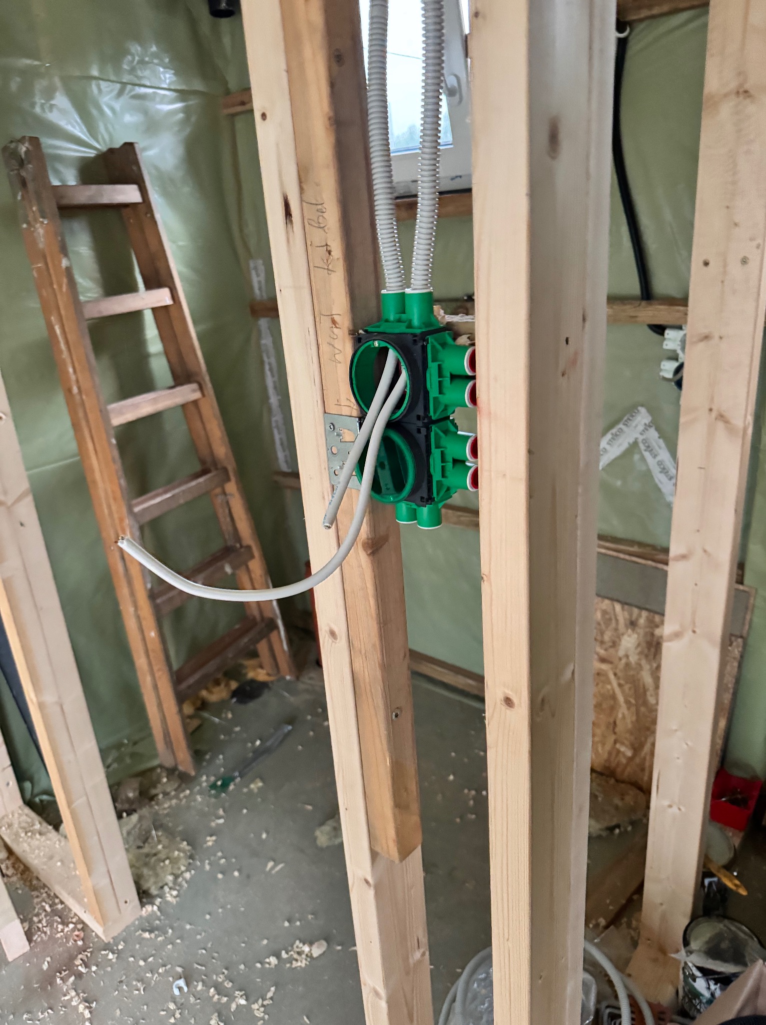 Instalacja wodna wewnątrz szkieletowej konstrukcji domu, z widoczną zieloną rozdzielnicą i przewodami. W tle drabina i folia izolacyjna.