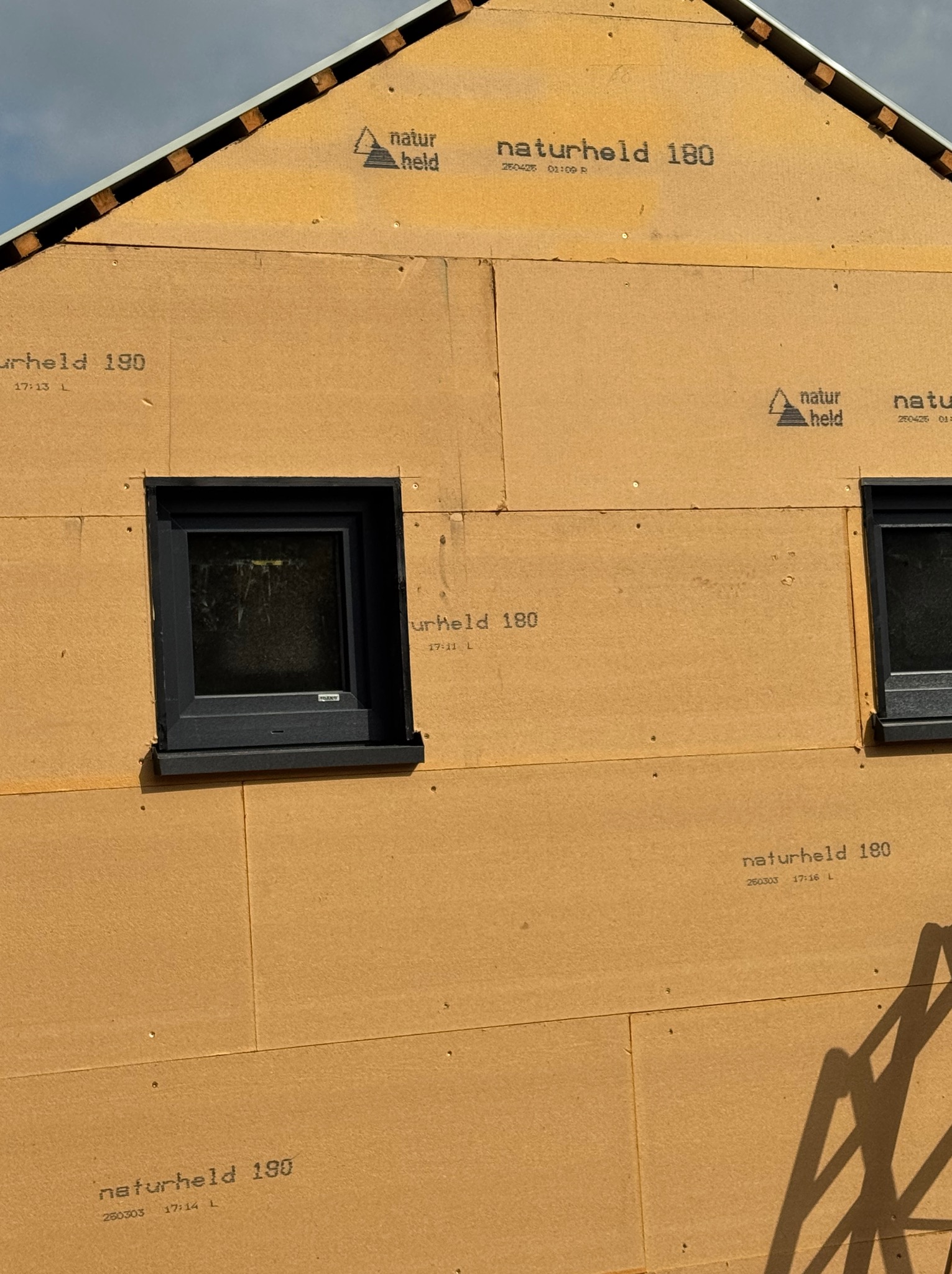 Fragment ściany domu szkieletowego w trakcie budowy, z widocznymi panelami izolacyjnymi i oknami. Widoczne oznaczenia paneli Naturheld 180 i 190.