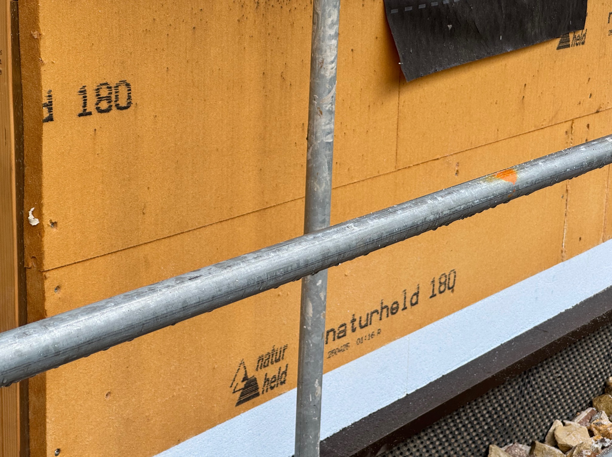 Fragment ściany domu w budowie, z widoczną izolacją 'naturheld 180', szarą rurą rusztowania i fragmentem czarnej membrany.