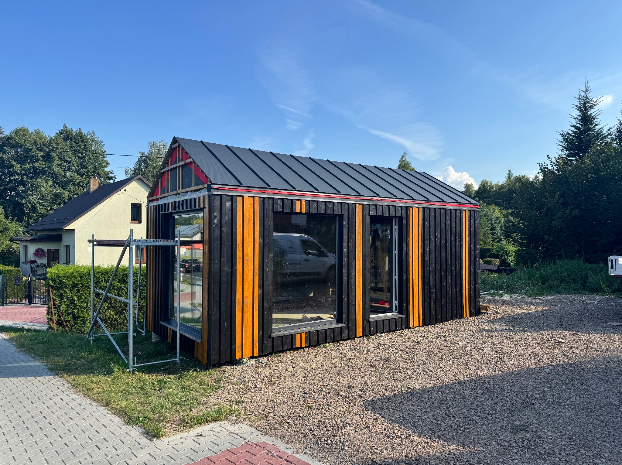 Częściowo ukończony, nowoczesny dom szkieletowy z czarnymi deskami i pomarańczowymi akcentami, widoczny z zewnątrz na tle błękitnego nieba i zieleni.