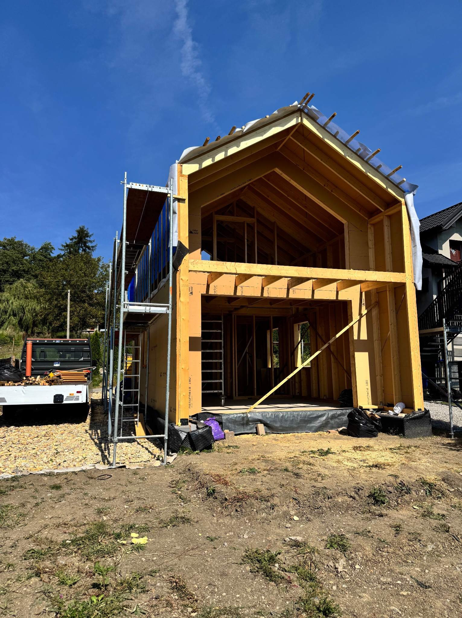 Wznoszenie dwukondygnacyjnego domu w konstrukcji szkieletowej. Widoczne drewniane elementy konstrukcyjne, rusztowania i materiały budowlane. Słoneczny dzień.