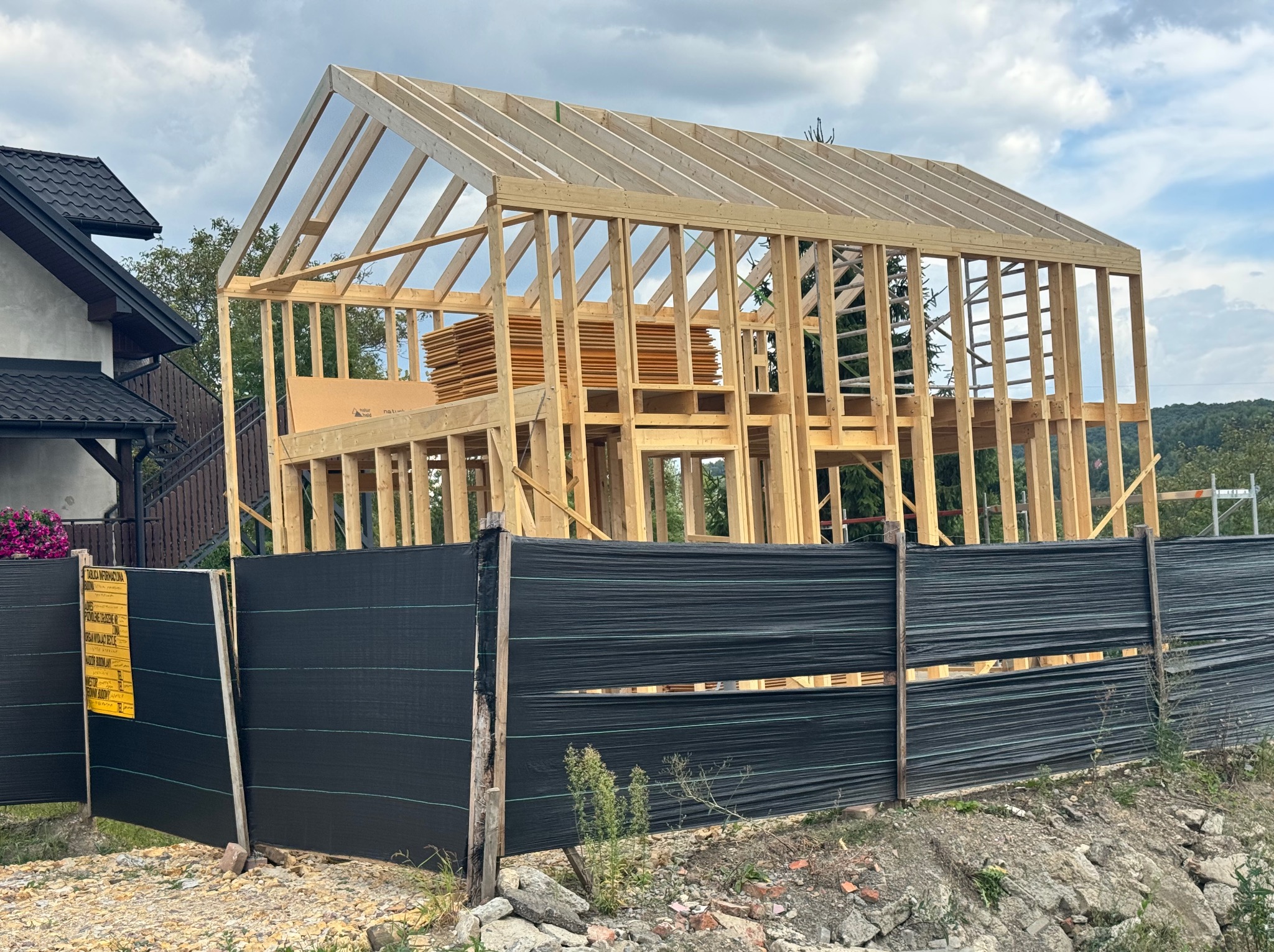 Konstrukcja szkieletowa domu w trakcie budowy, widok zza czarnego ogrodzenia budowlanego. Stos płyt OSB wewnątrz konstrukcji. Widoczne elementy dachu i ścian.