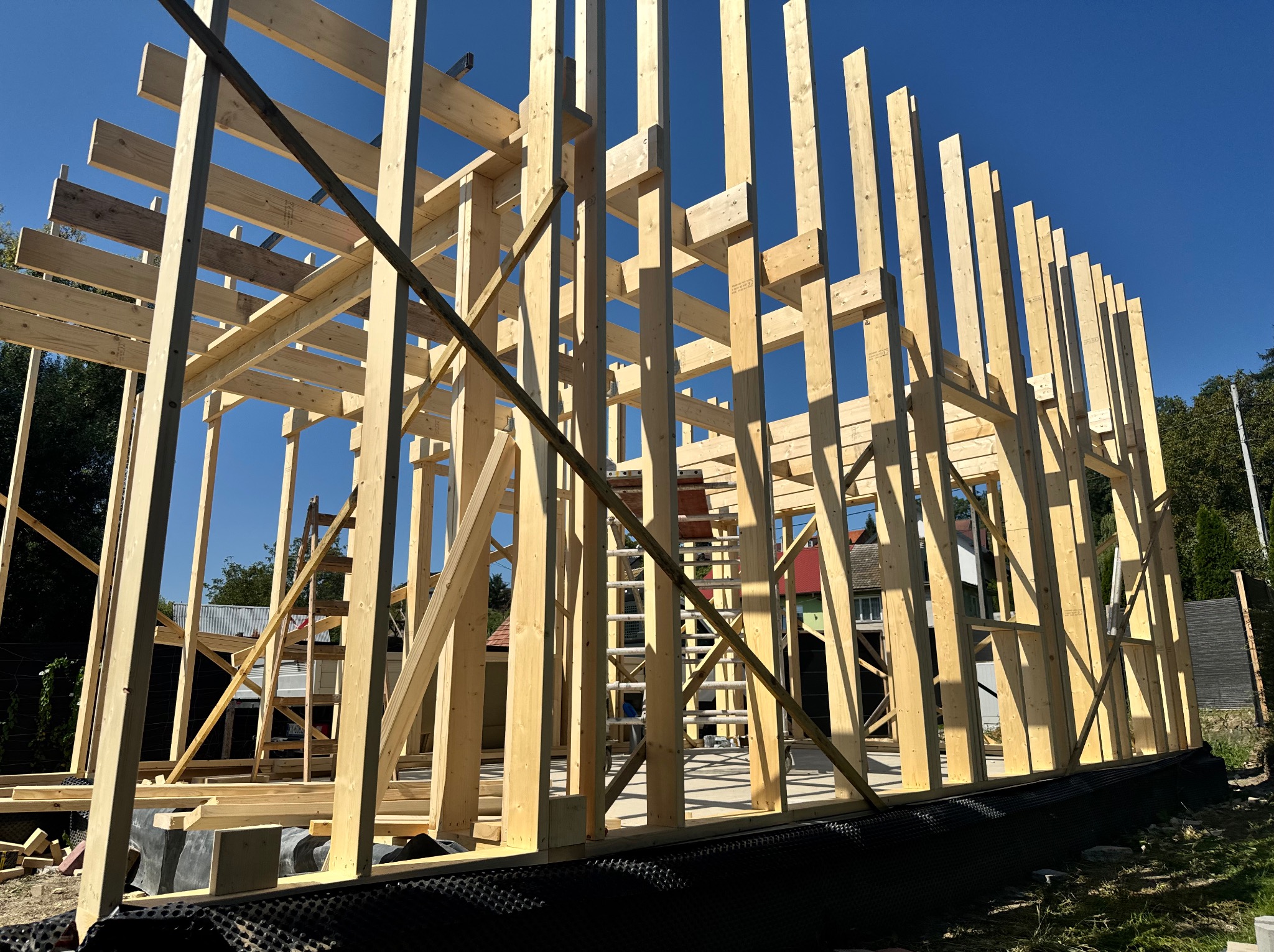 Konstrukcja szkieletowa domu w trakcie budowy, widok z zewnątrz, drewniane elementy na tle błękitnego nieba, drabina oparta o ścianę, widoczne sąsiednie budynki.