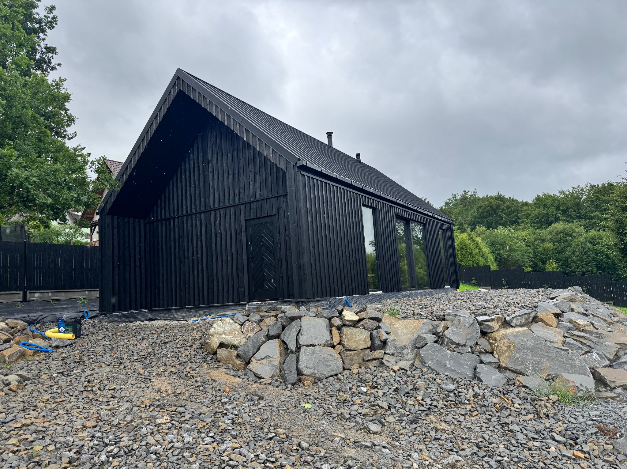 Czarny, nowoczesny dom z drewnianą fasadą i dwuspadowym dachem, otoczony kamiennym nasypem i zielenią, uchwycony w pochmurny dzień.