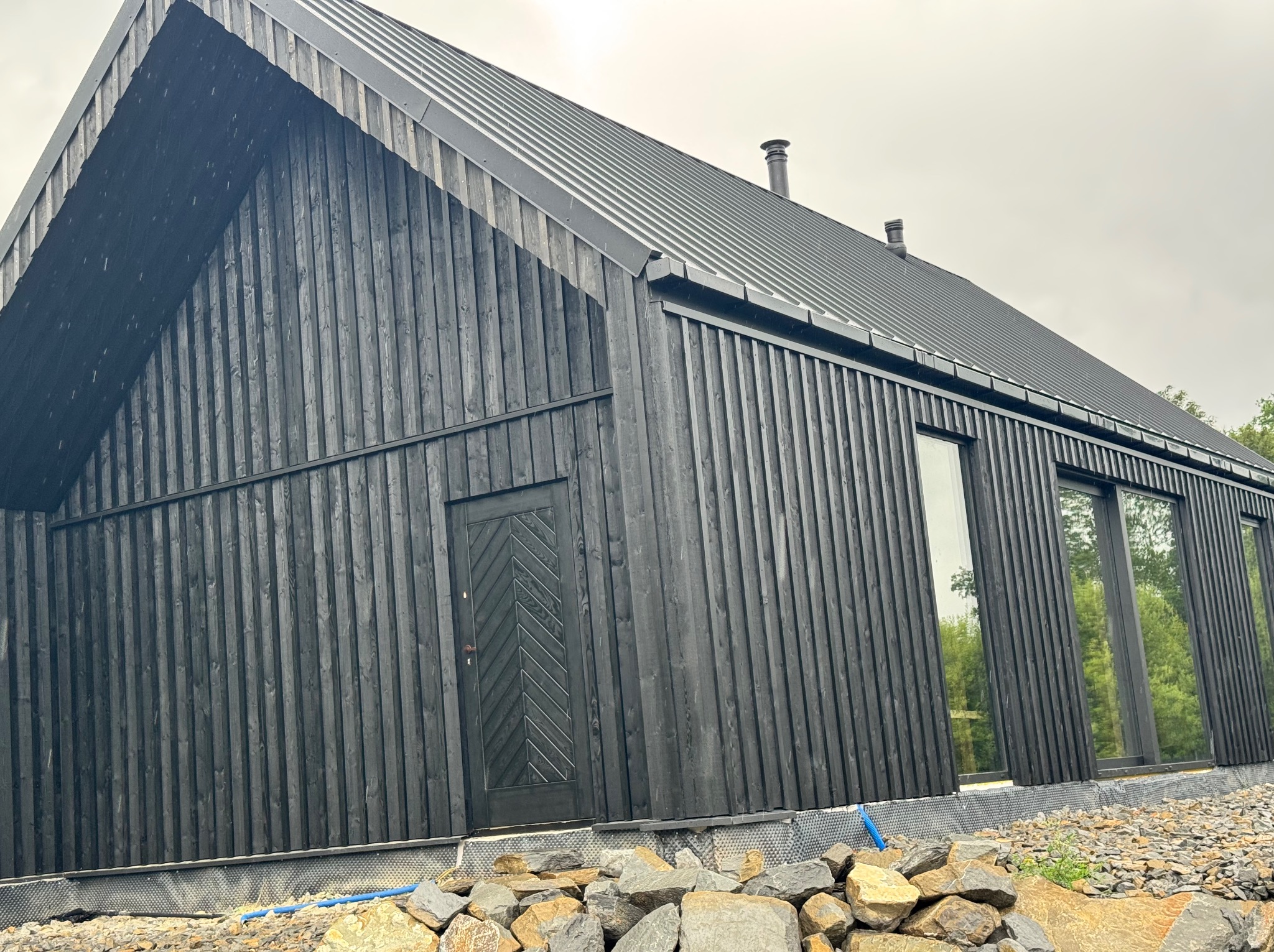 Nowoczesny, czarny dom z drewna o pionowej elewacji, z metalowym dachem i dużymi oknami. Uwagę przyciąga ciemna kolorystyka i minimalistyczny design. Częściowo widoczne fundamenty.