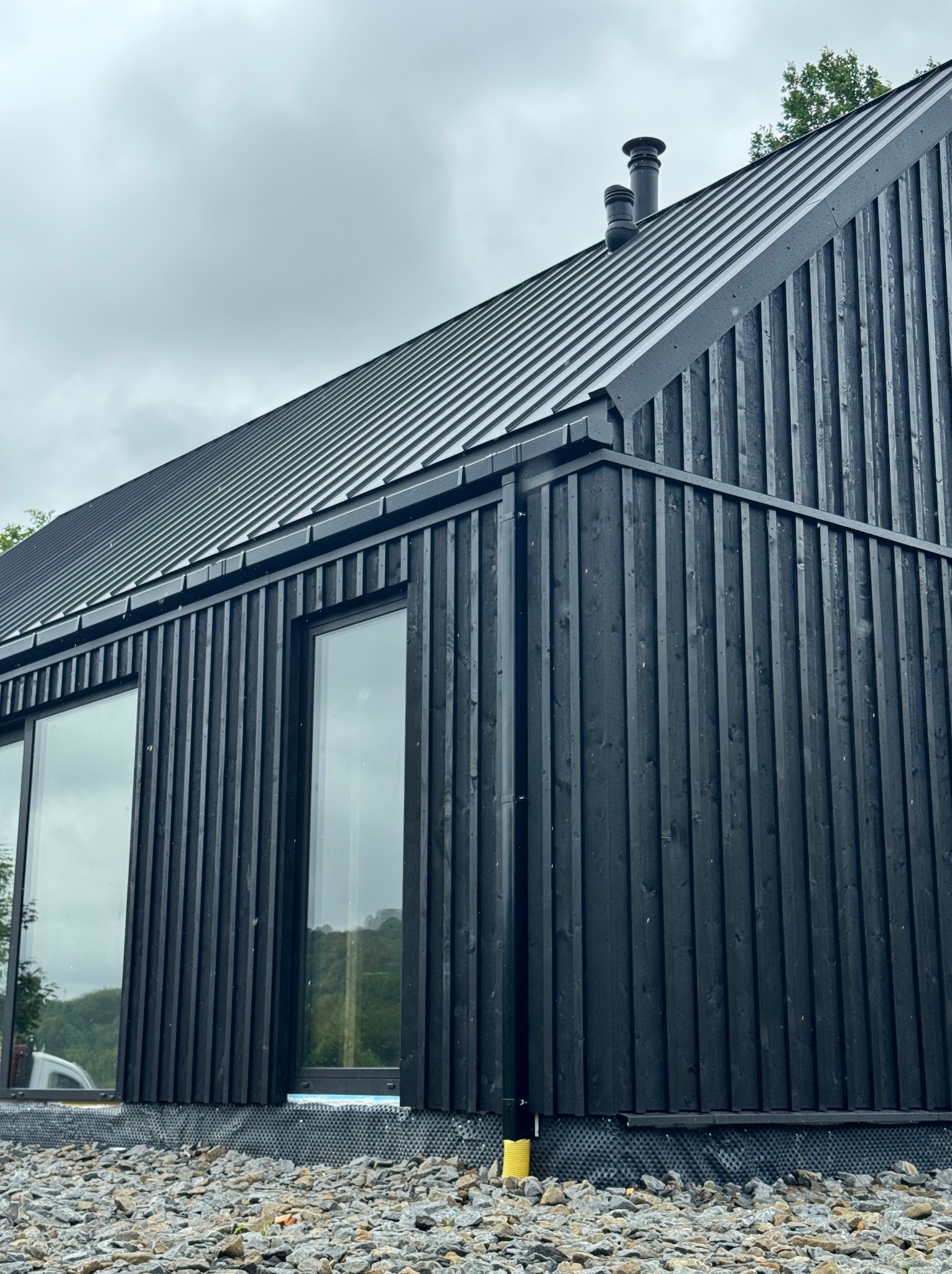 Czarny dom szkieletowy z metalowym dachem i dużymi oknami, wykończony pionowymi deskami. Widoczny fragment elewacji i otoczenie z kamieni.