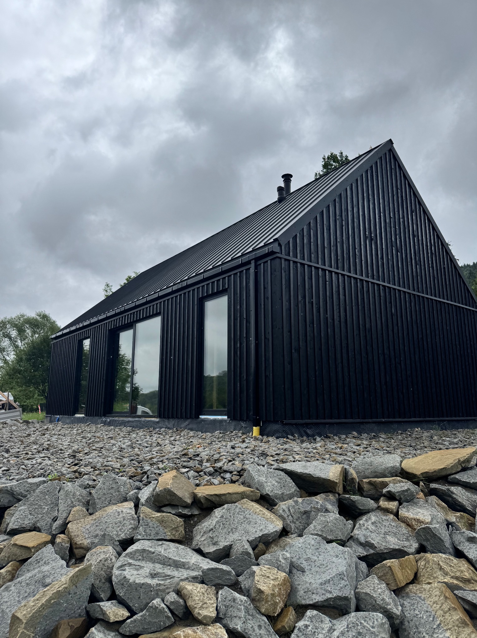 Nowoczesny, czarny dom z drewna o minimalistycznej architekturze, z dużymi oknami i metalowym dachem, otoczony kamieniami. Pochmurne niebo w tle.