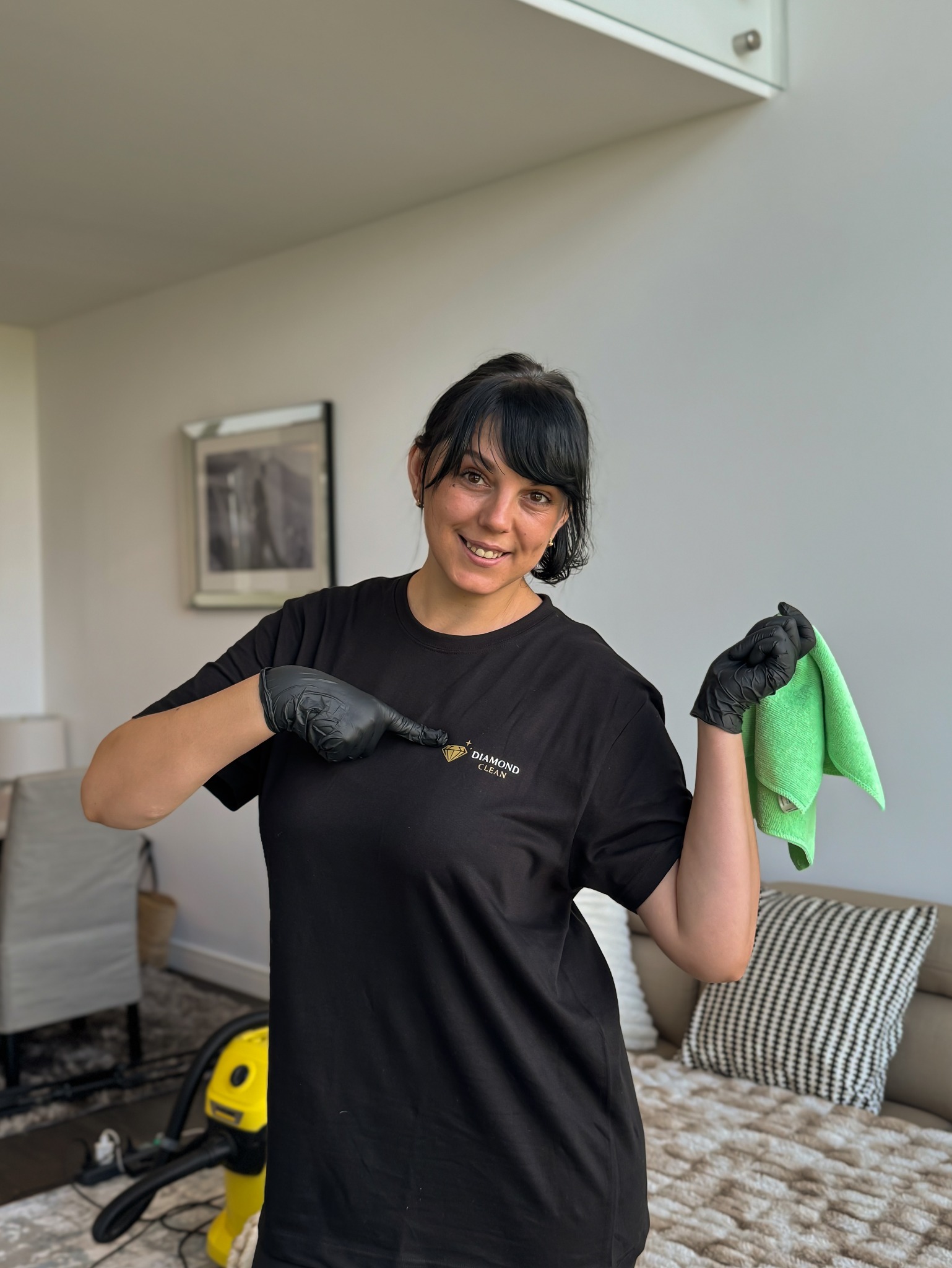 Uśmiechnięta kobieta w czarnej koszulce z logo 'Diamond Clean' wskazuje na nie, trzymając zieloną ściereczkę. W tle odkurzacz i wnętrze mieszkania.