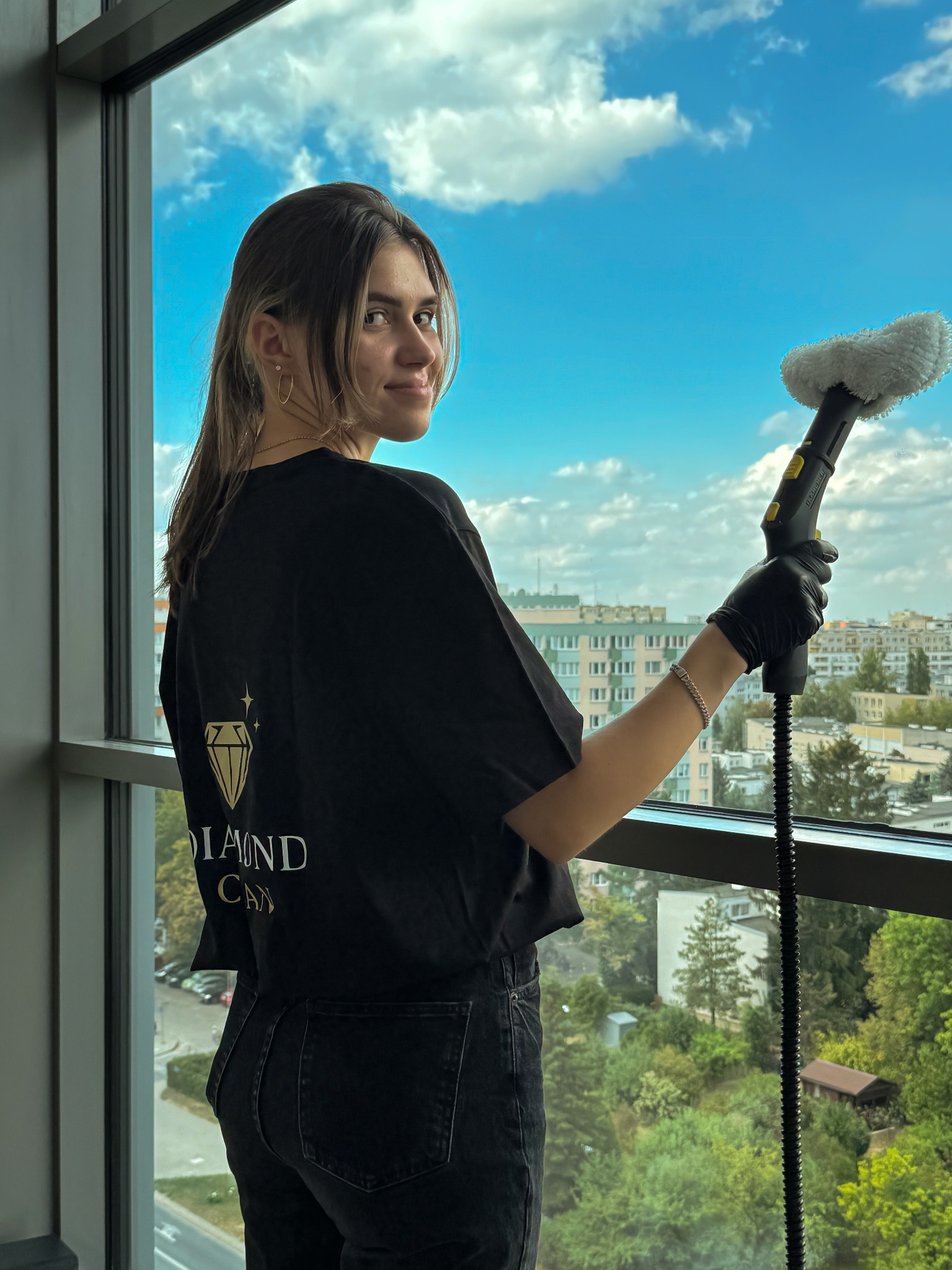 Kobieta w czarnej koszulce z logo Diamond Clean myje okno za pomocą parownicy Karcher, widok na miasto w tle.