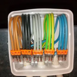 Zestawienie kolorowych przewod&oacute;w w białej puszce instalacyjnej, połączonych pomarańczowymi złączkami WAGO. Widok z g&oacute;ry na uporządkowane okablowanie.