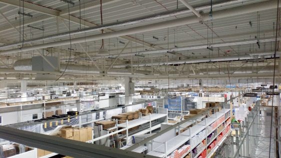 Rozległa hala magazynowa z widocznymi instalacjami pod sufitem, rurami wentylacyjnymi i kablami. Oświetlenie sufitowe równomiernie rozproszone po całej przestrzeni.