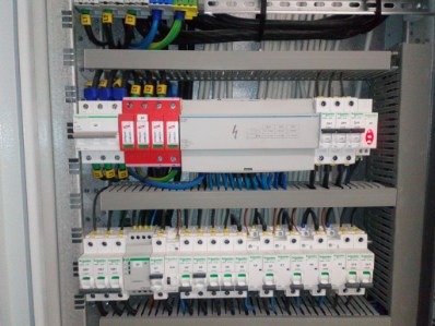 Rozdzielnica elektryczna z zabezpieczeniami przepięciowymi i wyłącznikami różnicowoprądowymi, widoczne kable i szyny montażowe.