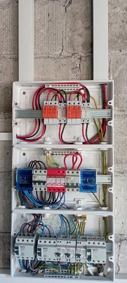 Otwarta, trzyczęściowa skrzynka elektryczna z zabezpieczeniami przepięciowymi, bezpiecznikami i okablowaniem na tle szarej ściany z cegieł.