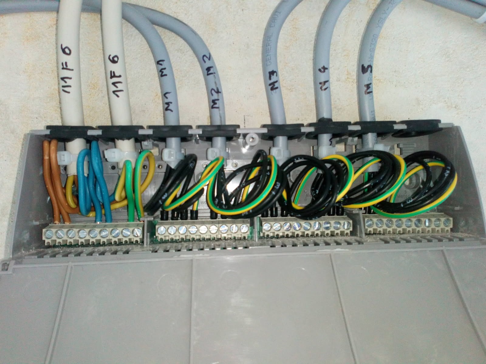 Szara skrzynka elektryczna z podłączonymi przewodami o różnych kolorach, oznaczonymi numerami. Widoczne złączki i kable wchodzące do skrzynki.