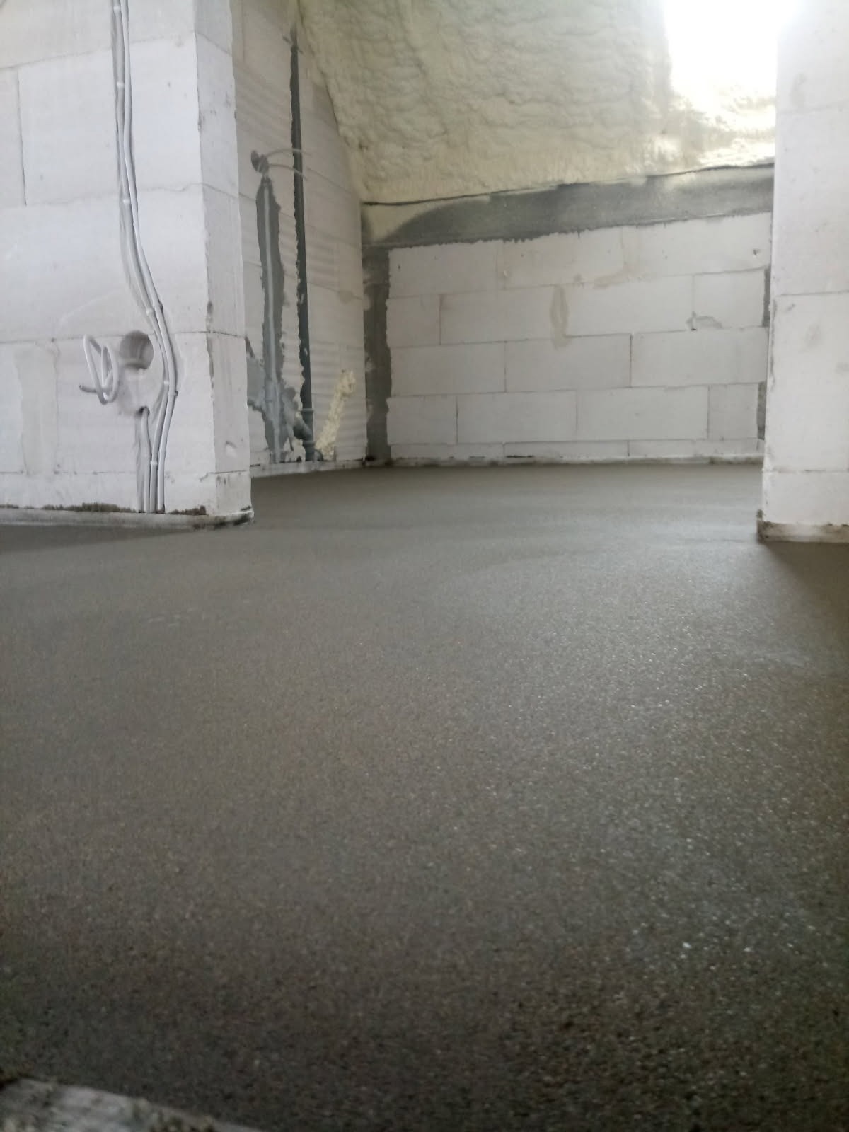 Wnętrze w trakcie prac wykończeniowych: wylana, świeża posadzka betonowa, ściany z białych bloczków z widocznymi instalacjami elektrycznymi. Poddasze ocieplone pianą.