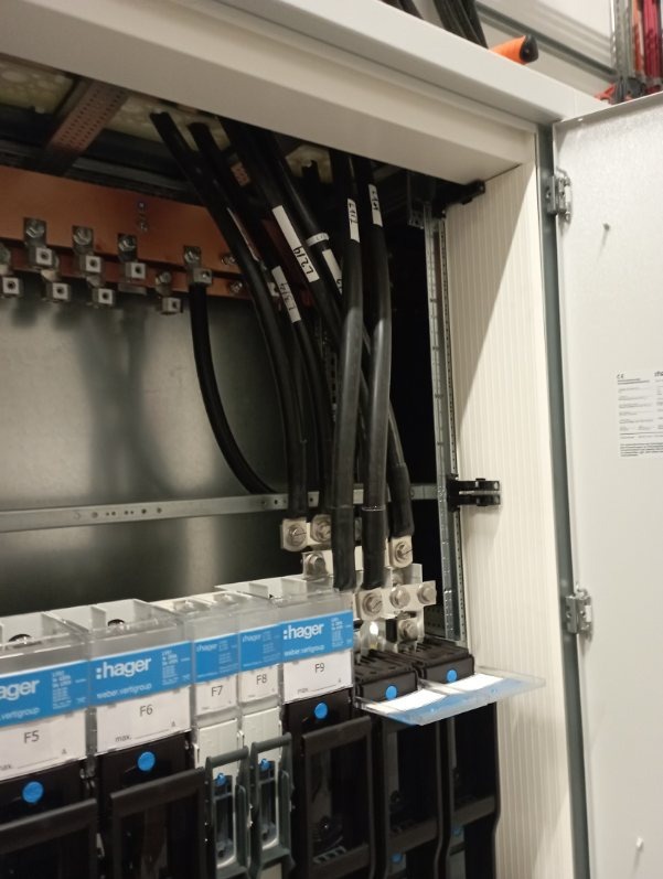 Wnętrze szafy elektrycznej z czarnymi przewodami oznaczonymi etykietami, podłączonymi do listwy zaciskowej i zabezpieczeń Hager. Widoczne elementy instalacji.