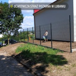 Łukasz Kerlin - Montaż panelowego ogrodzenia posesji w Nowej Karczmie. Widoczny fragment budynku i osoba z wiadrem wzdłuż linii ogrodzenia. Trawa i drzewa w tle.
