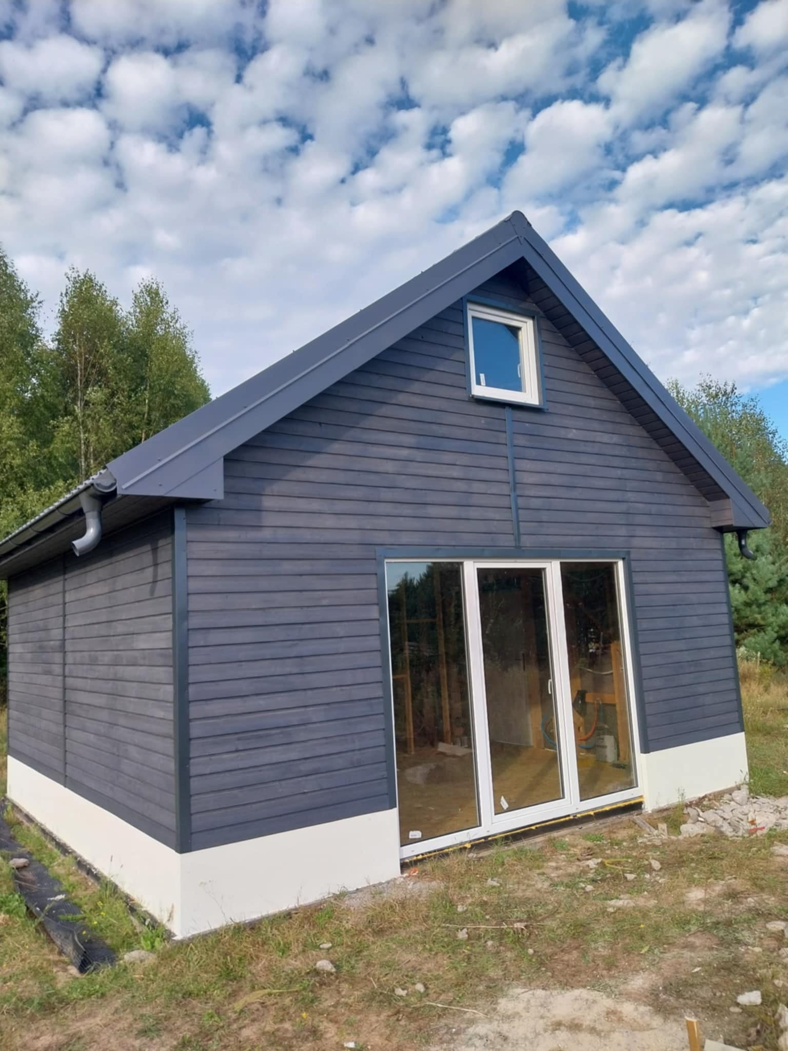 Nowoczesny, szary domek narzędziowy z białym fundamentem i dużymi, przesuwnymi drzwiami. Widok na wnętrze w stanie surowym, błękitne niebo z chmurami w tle.