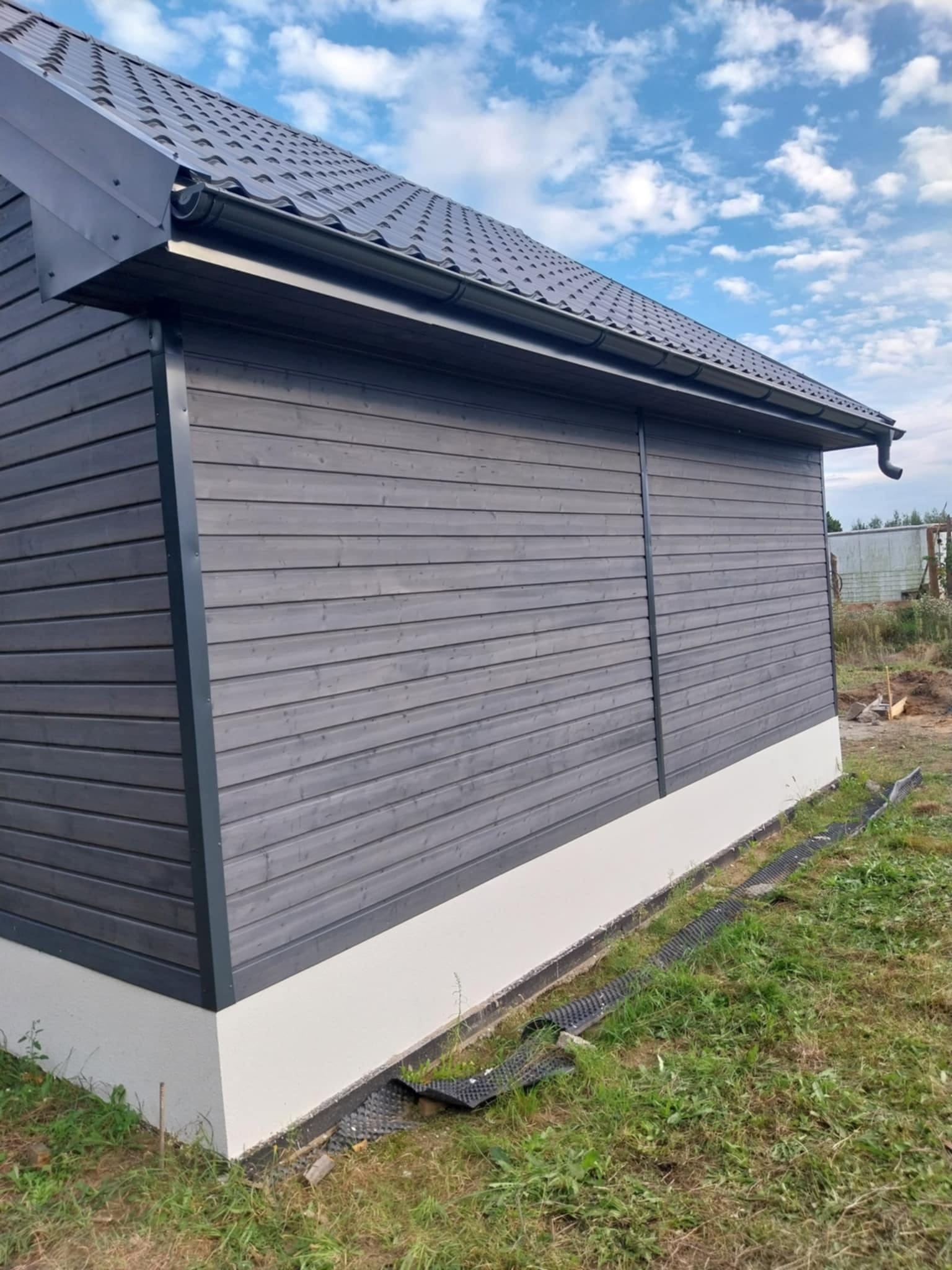 Szary budynek z drewnianą elewacją i metalowym dachem, widok z boku na tle błękitnego nieba z chmurami. Widoczne rynny i fragment fundamentu.