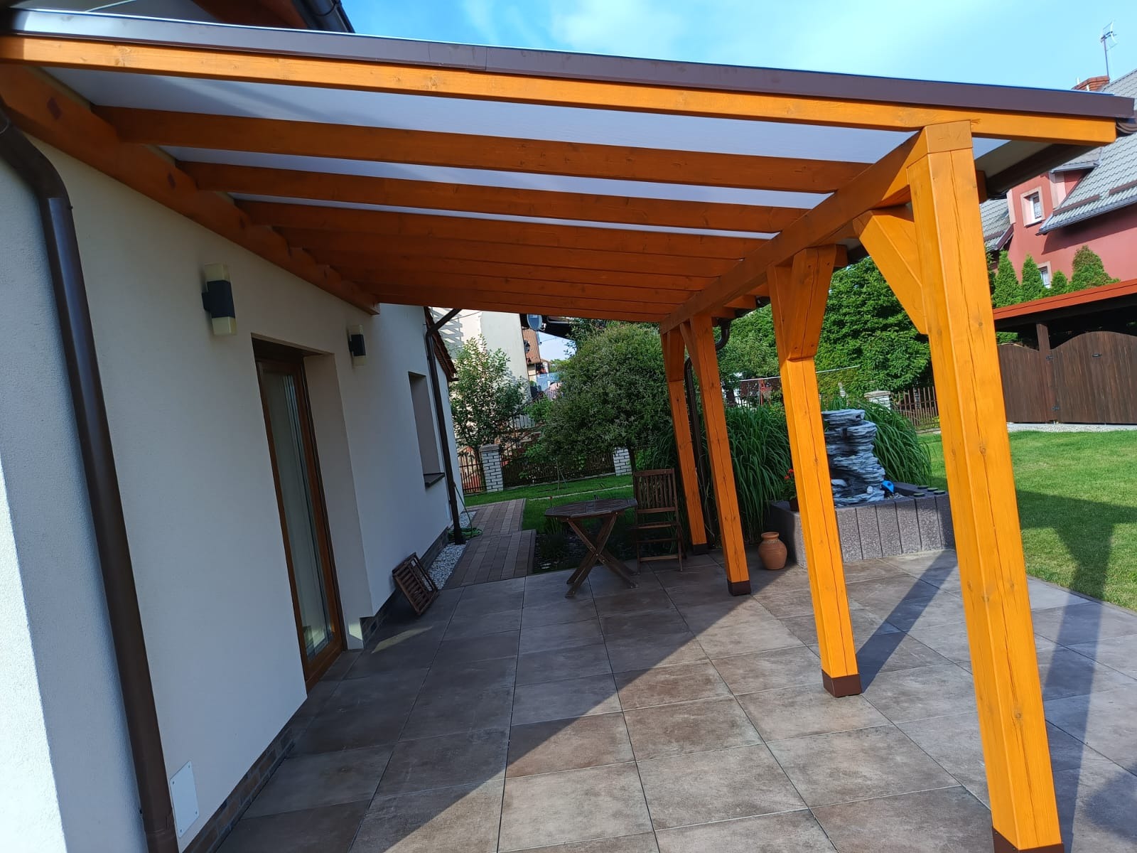 Zadaszona, drewniana pergola z przezroczystym dachem, przylegająca do domu. Widoczny stół z krzesłem i fragment ogrodu. Ułożona kostka brukowa na tarasie.