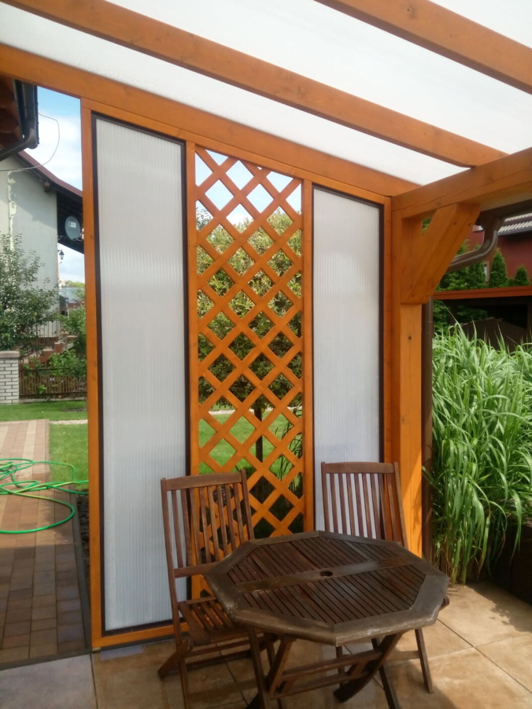 Drewniana pergola z ażurowym panelem i przezroczystymi ściankami, stolik z krzesłami na tarasie. Widok na ogród przez kratkę.