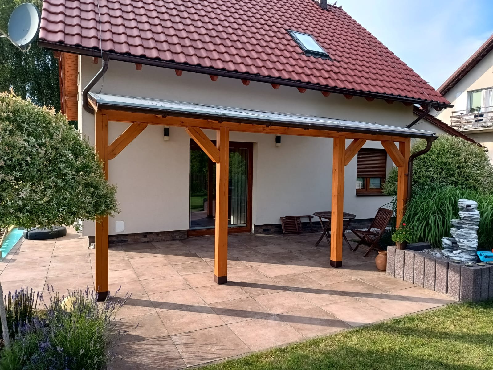 Drewniana pergola z przezroczystym zadaszeniem przylegająca do domu, z widoczną kostką brukową, roślinnością i elementami dekoracyjnymi ogrodu.