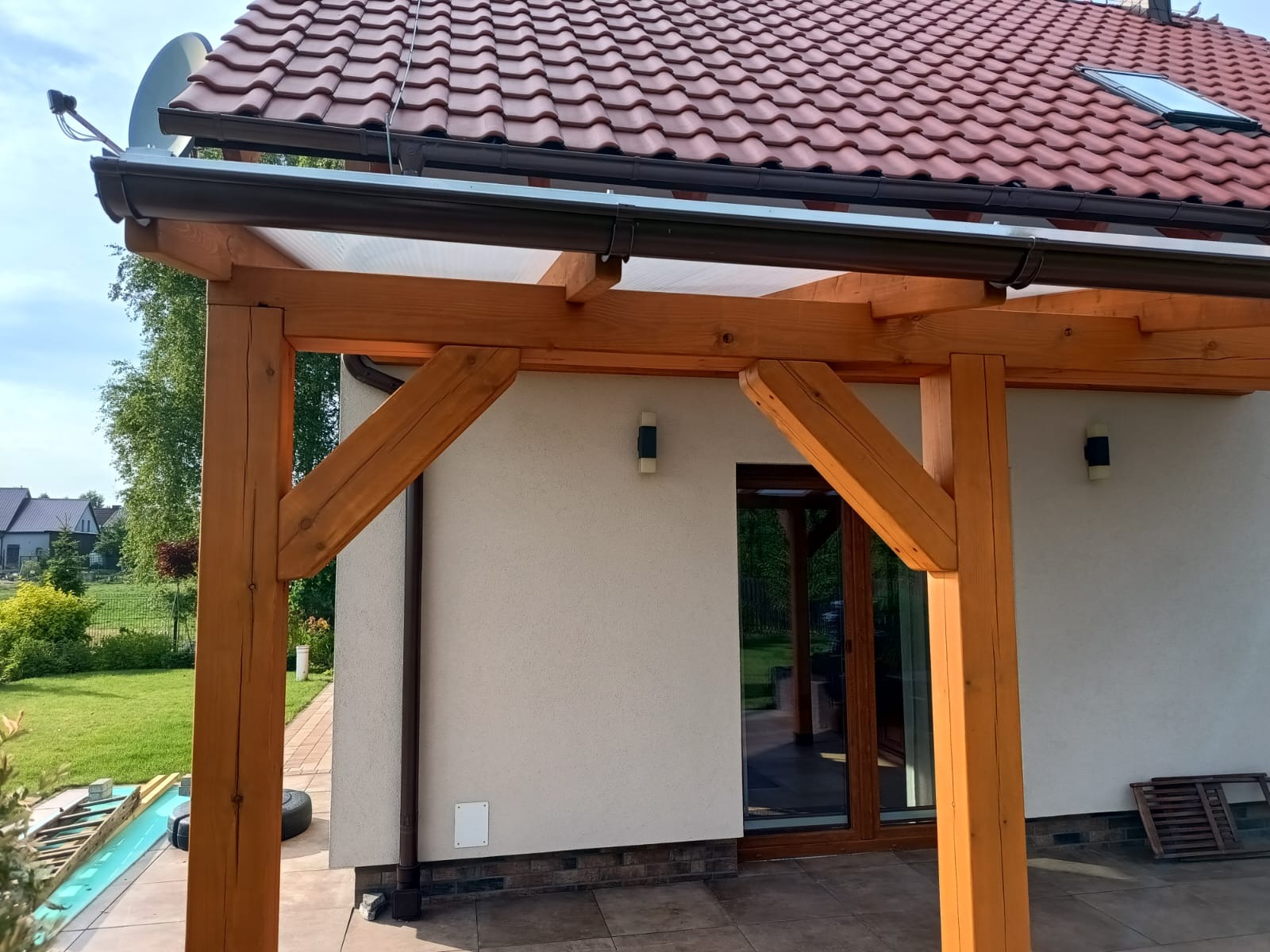 Drewniana pergola przy domu z brązową dachówką, widok z boku. Elementy konstrukcyjne z belek, widoczne detale łączeń. Tło: ściana budynku i fragment ogrodu.