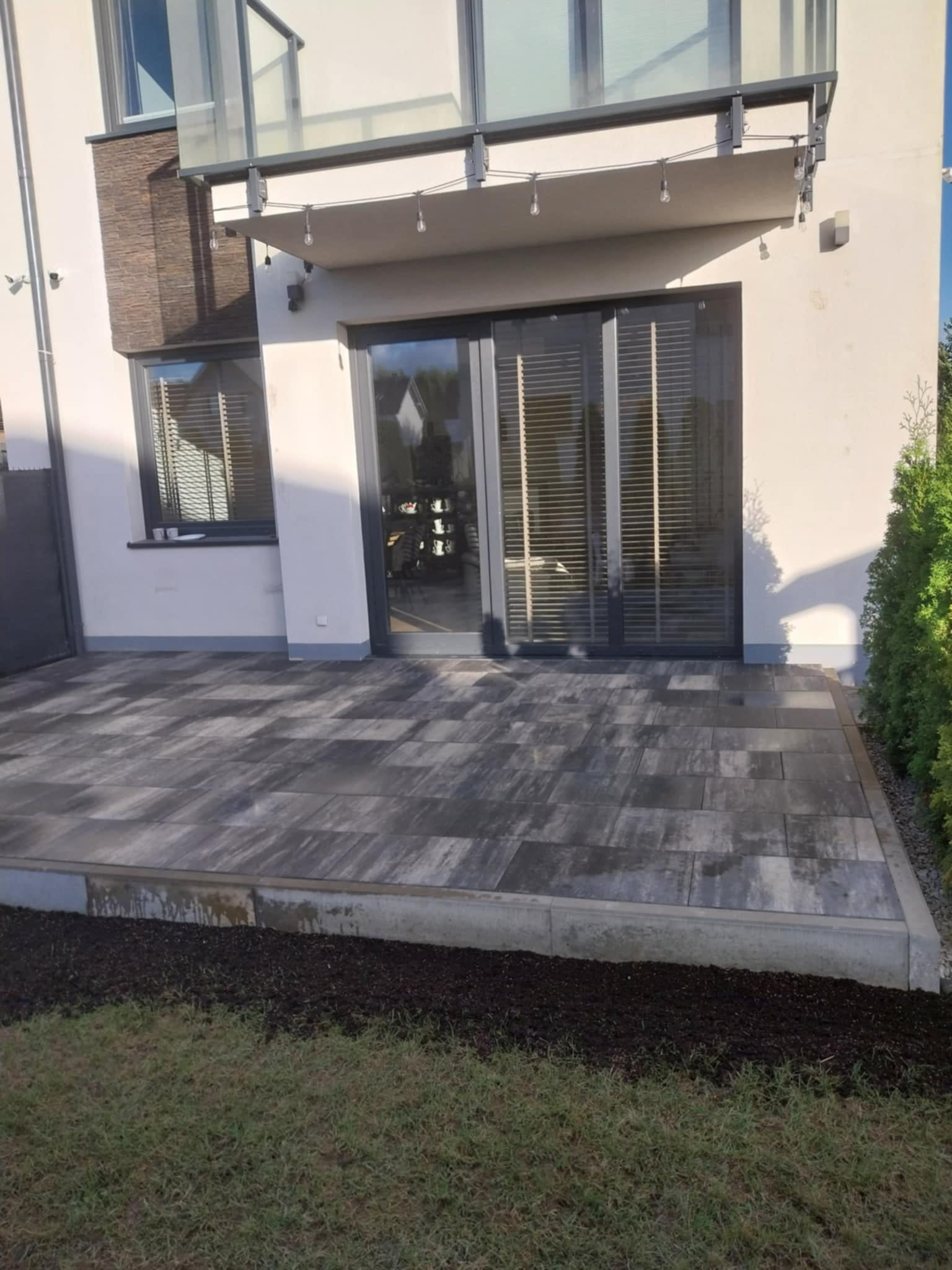 Nowoczesny taras z szarych płyt brukowych, zadaszony balkon z oświetleniem, widok na dom z dużymi oknami i zielony trawnik. Minimalistyczny design.