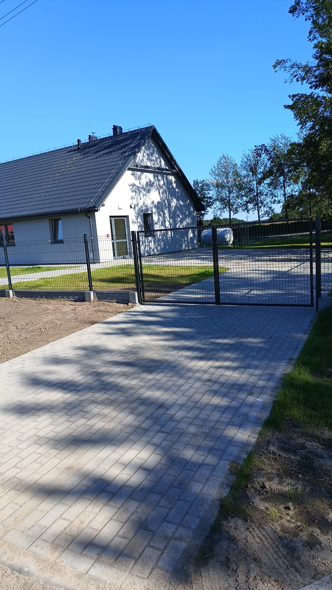 Nowoczesny dom z szarą kostką brukową na podjeździe i czarnym ogrodzeniem panelowym z bramą wjazdową. Widok na budynek i zadbany trawnik.