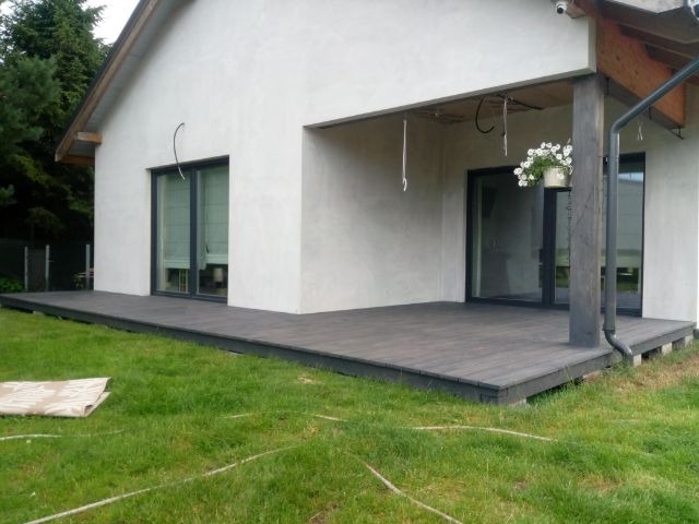 Nowoczesny taras z desek kompozytowych przy białym domu z dużymi oknami. Donica z kwiatami pod dachem. Widoczne elementy konstrukcyjne tarasu.