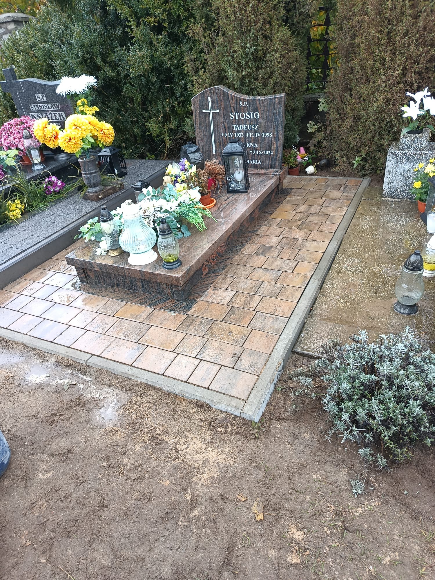 Ułożona kostka brukowa wokół nagrobka z kwiatami i zniczami. Nowa nawierzchnia z kostki w odcieniach brązu. Widok na cmentarz.