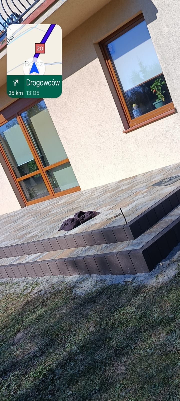 Schody wejściowe wyłożone kostką brukową w odcieniach szarości i brązu, z widocznym fragmentem elewacji budynku i trawnika. Na kostce leży ciemna bluza.