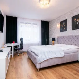 Nowoczesna sypialnia z łóżkiem typu king-size, biurkiem i telewizorem. Tapeta w kwiaty, ciemne zasłony, drewniana podłoga. Oświetlenie sufitowe i lampy nocne.