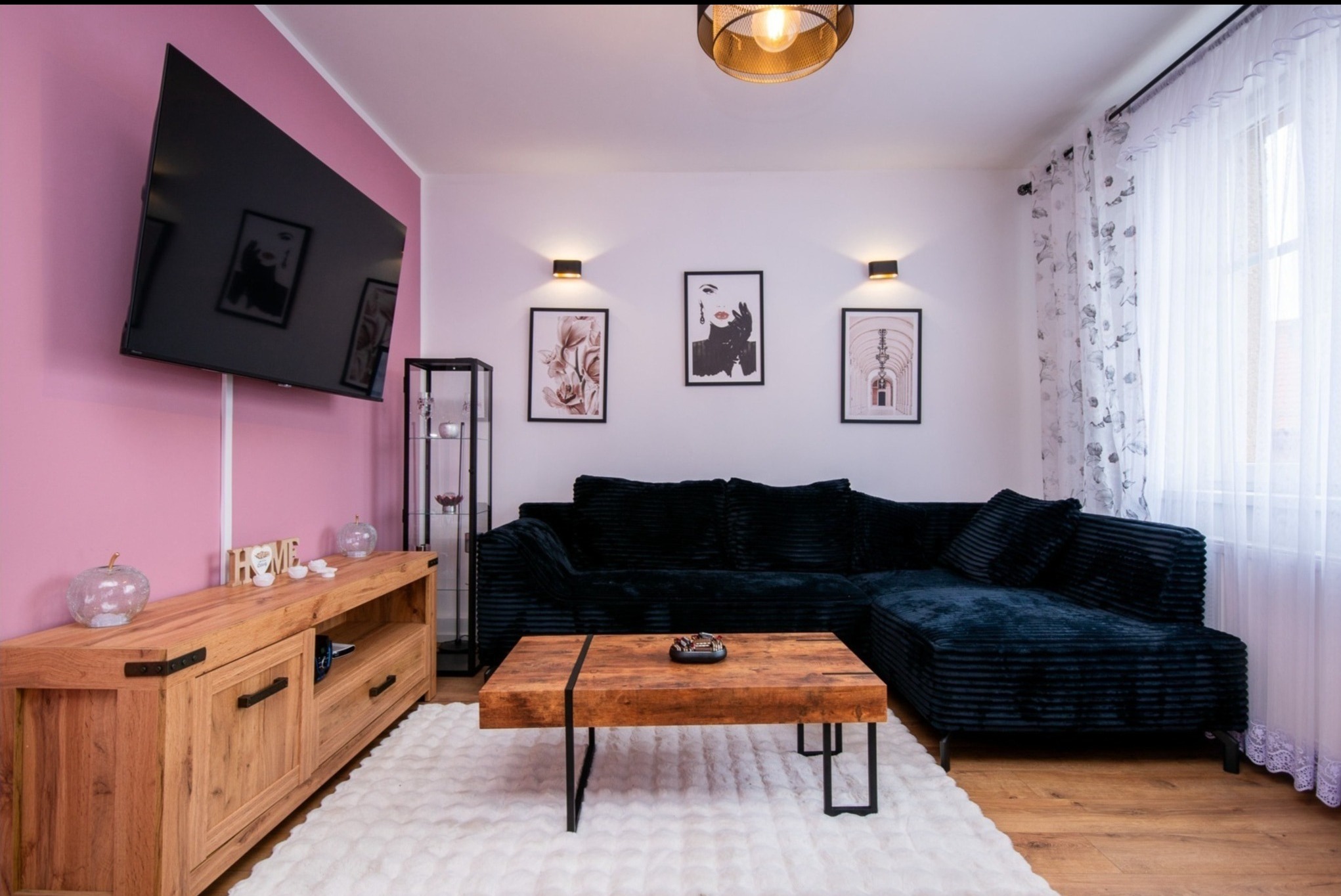 Salon z czarną sofą narożną, drewnianym stolikiem kawowym i komodą. Ściana w kolorze pudrowego różu. Nowoczesne oświetlenie i dekoracje.