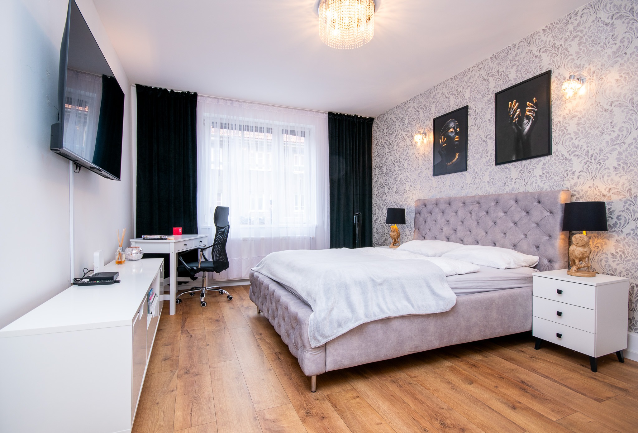 Nowoczesna sypialnia z łóżkiem typu king-size, biurkiem i telewizorem. Tapeta w kwiaty, ciemne zasłony, drewniana podłoga. Oświetlenie sufitowe i lampy nocne.