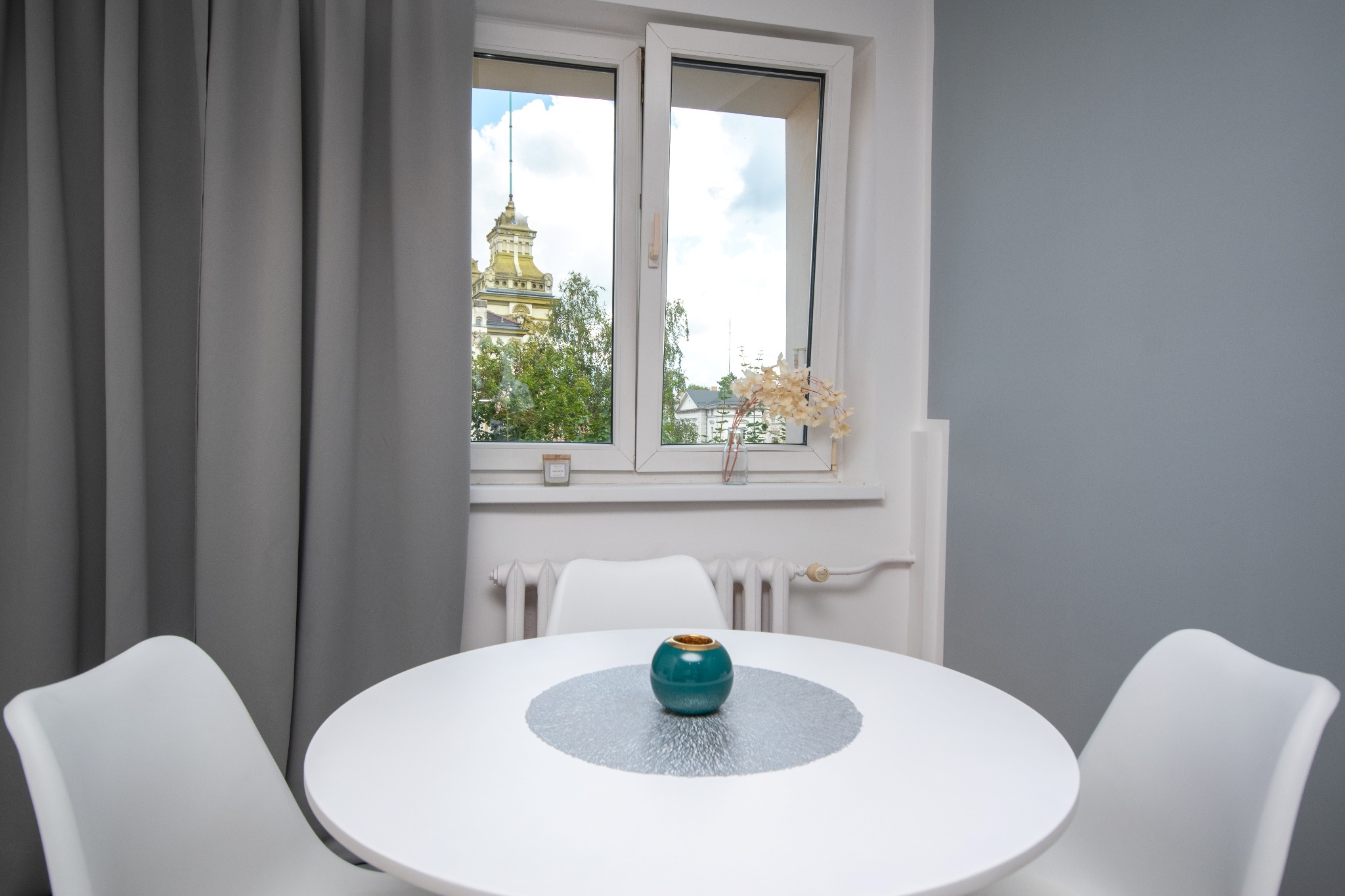 Jasne wnętrze: biały okrągły stół z dekoracją, trzy krzesła. Za oknem widok na budynek z wieżą. Szare zasłony i ściana w tle.
