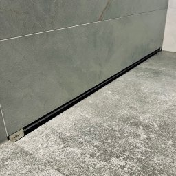 INWELL INSTALACJE MICHAŁ KUBICA - Szczegółowe ujęcie liniowego odpływu prysznicowego Viega, wkomponowanego w szarą, kamienną posadzkę i ścianę z płytek o podobnej fakturze. Minimalistyczny design.