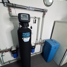 INWELL INSTALACJE MICHAŁ KUBICA - Instalacja uzdatniania wody Water Technic z lampą UV, rurami i niebieskim zbiornikiem. Widoczne elementy sterujące i logo Inwell. Szare płytki podłogowe.