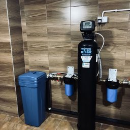 INWELL INSTALACJE MICHAŁ KUBICA - Czarny system uzdatniania wody Water Technic Pro Multi z niebieskim zbiornikiem na solankę, zamontowany w pomieszczeniu wyłożonym płytkami imitującymi drewno.