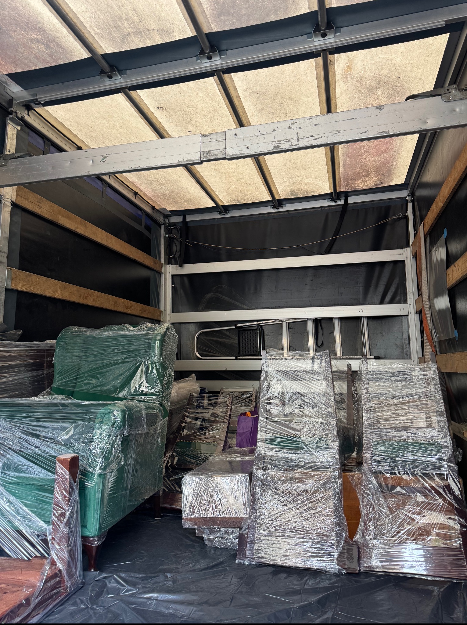 Wnętrze przestrzeni ładunkowej busa wypełnione meblami zabezpieczonymi folią stretch, przygotowanymi do transportu. Widoczna drabina i elementy konstrukcyjne pojazdu.