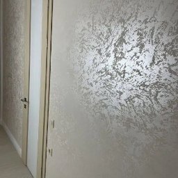 Reco bud - Ściana z dekoracyjną, srebrzystą fakturą obok kremowych drzwi z ościeżnicą. Widoczne gniazdka elektryczne. Całość w jasnej tonacji, z delikatnym wzorem na ścianie.