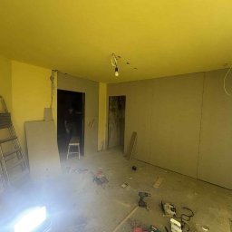 Reco bud - Pokój w trakcie remontu: ściany z płyt karton-gips, widoczne narzędzia, drabina oparta o ścianę. Żółte ściany i sufit. W tle widoczna postać w przejściu.