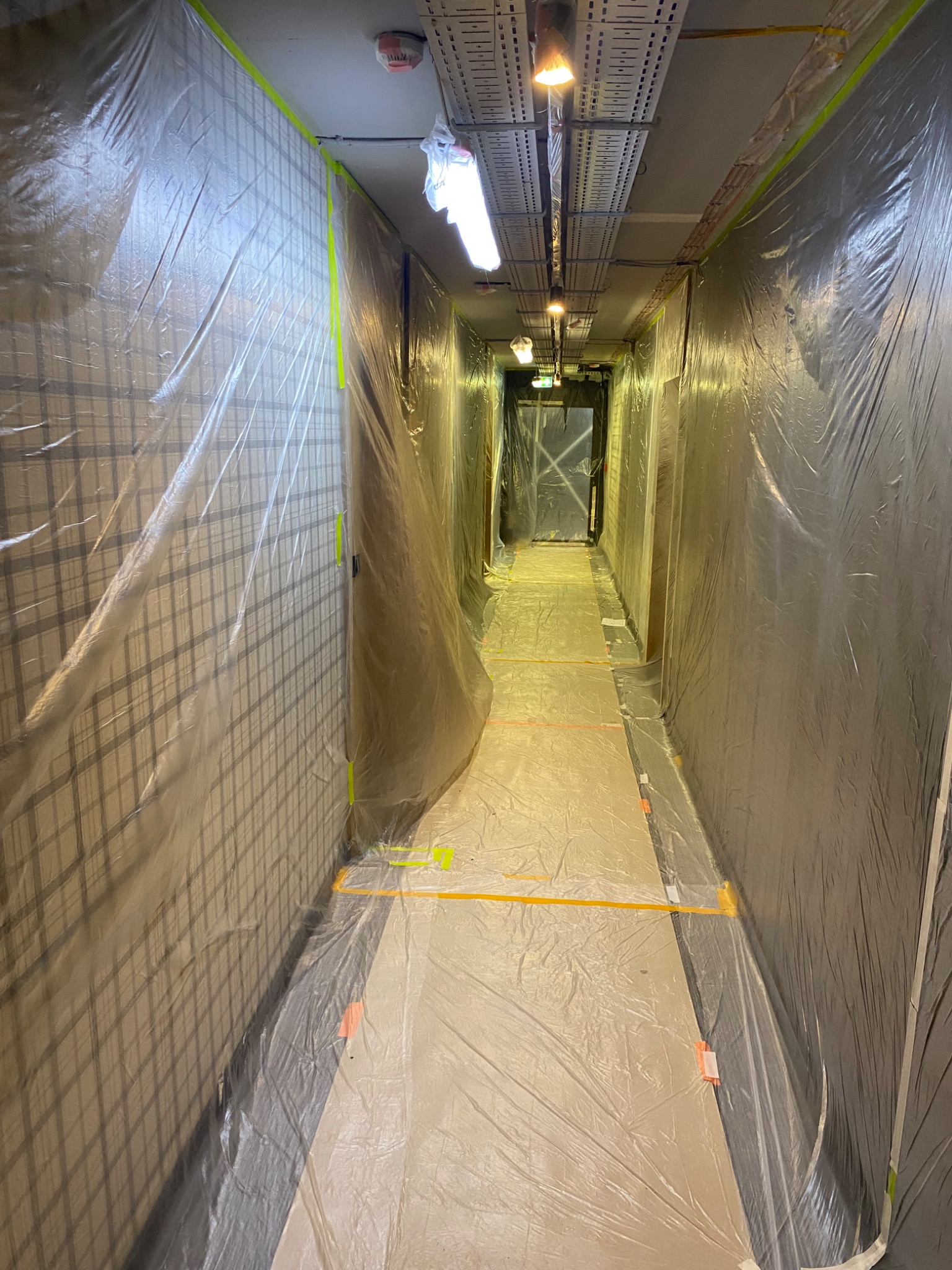 Korytarz zabezpieczony folią malarską i taśmą przed pracami wykończeniowymi. Widoczne oświetlenie sufitowe i instalacje. Przygotowanie do malowania.