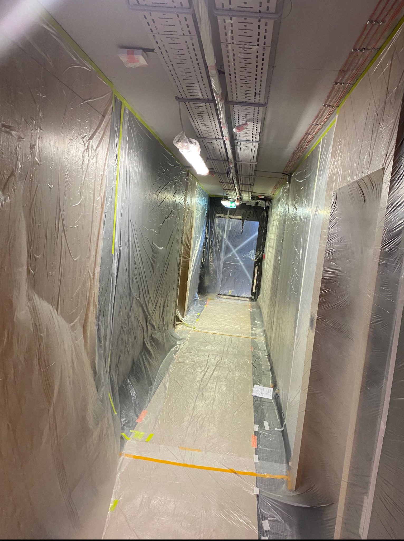 Korytarz zabezpieczony folią i taśmą malarską przed pracami remontowymi. Widoczne instalacje pod sufitem. Przygotowanie do malowania lub szpachlowania.