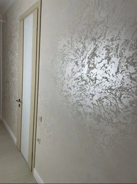 Ściana z dekoracyjną, srebrzystą fakturą obok kremowych drzwi z ościeżnicą. Widoczne gniazdka elektryczne. Całość w jasnej tonacji, z delikatnym wzorem na ścianie.