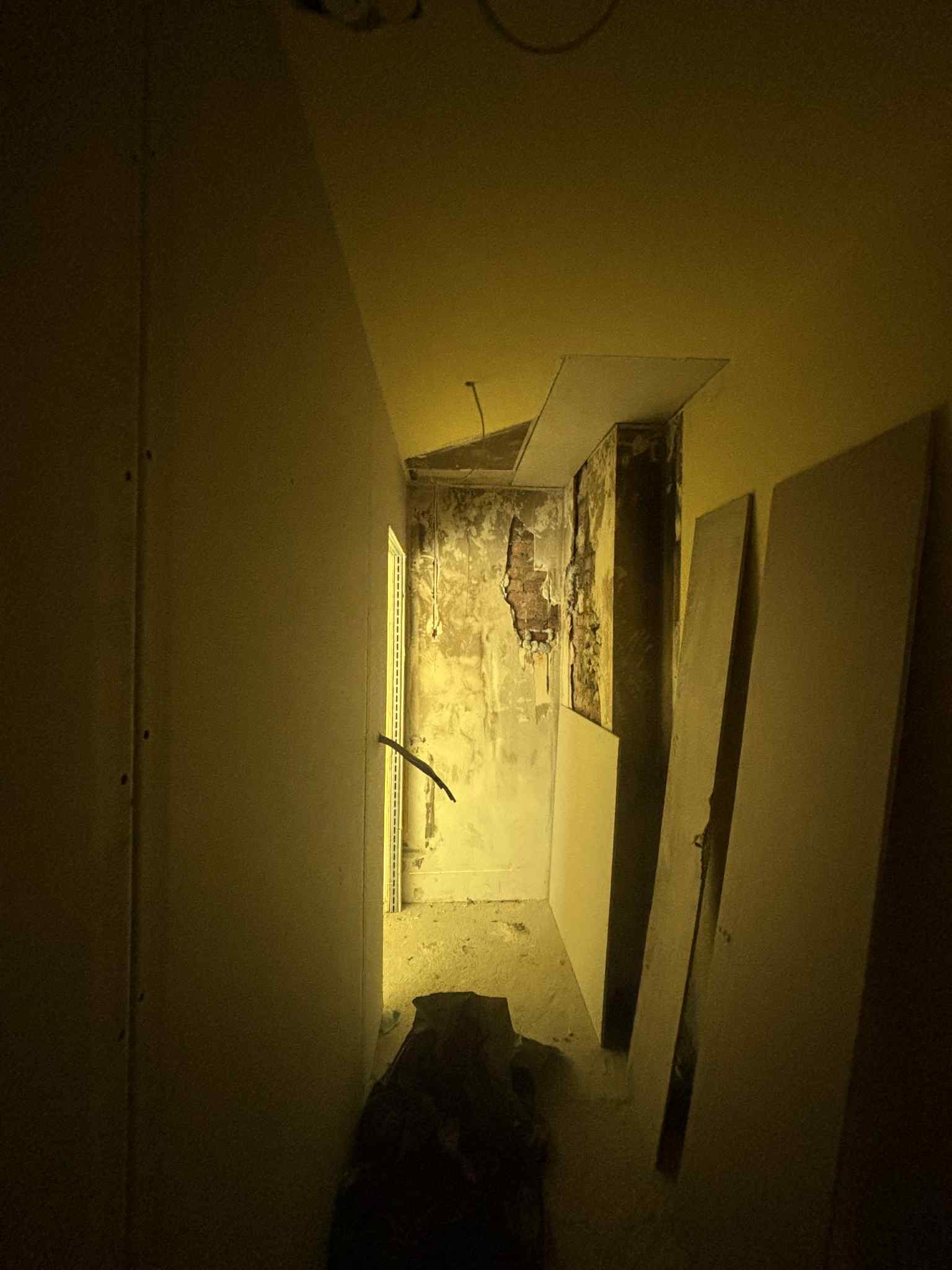 Korytarz w trakcie remontu: odsłonięta cegła, płyty karton-gips oparte o ścianę, widoczne instalacje. Przyciemnione światło, surowy stan wykończenia.